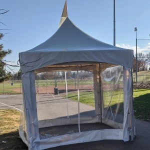 Gray peaked event tent with clear mesh windows sits on pavement near grassy area.