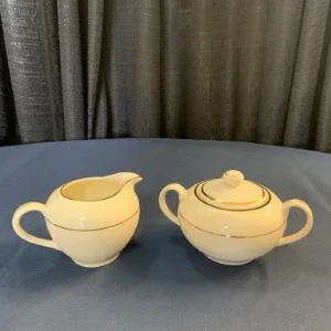 Soft yellow ceramic cream and sugar set with curved handles on blue tablecloth near curtain.
