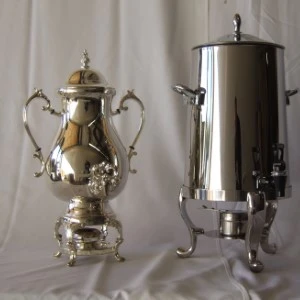 Silver samovar and ornate coffee urn with decorative handles on white draped fabric.