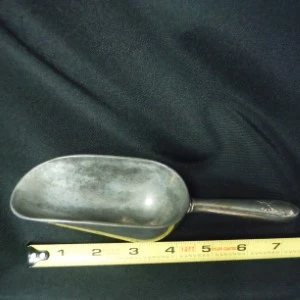 Vintage aluminum metal scoop with worn handle, measured against yellow measuring tape on dark background.