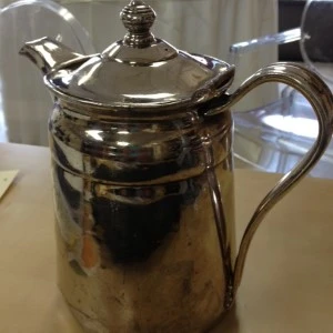 Silver metallic vintage pitcher with ornate lid and curved handle, reflecting light.