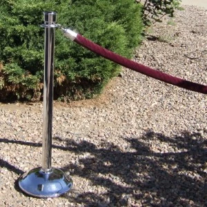 Stainless steel stanchion post with burgundy velvet rope on gravel ground near shrubs.