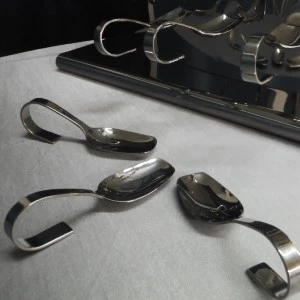 Silver curved spoons with bent handles rest on a white surface near a dark reflective background.