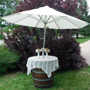 White market umbrella mounted on wooden barrel with lace tablecloth in garden setting.