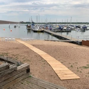 Wooden boardwalk leads to sandy beach with marina dock and sailboats in background.