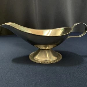 Gold metallic gravy boat with curved handle sits on pedestal base against dark blue background.