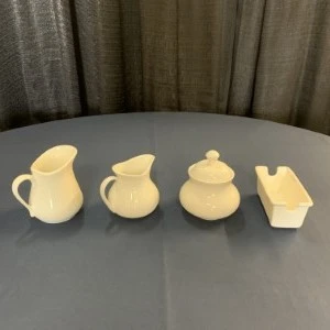 Four cream-colored ceramic serving pieces including pitcher, sugar bowl, and rectangular dish on gray tablecloth.