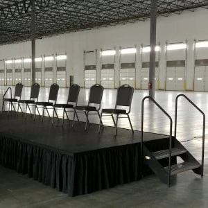 Black stage platform with black skirting, metal chairs, and side stairs in industrial event space with white walls.