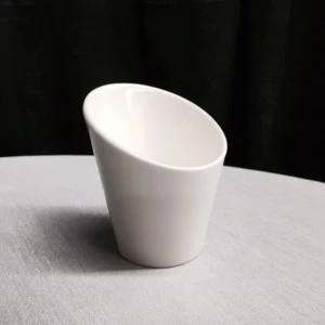White ceramic angled cup tilts on textured gray tablecloth against dark background.